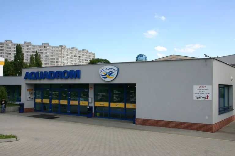 Šedá přízemní budova Aquadromu Most s modrým nápisem Aquadrom a prosklenými vstupními dveřmi, v pozadí panelový dům