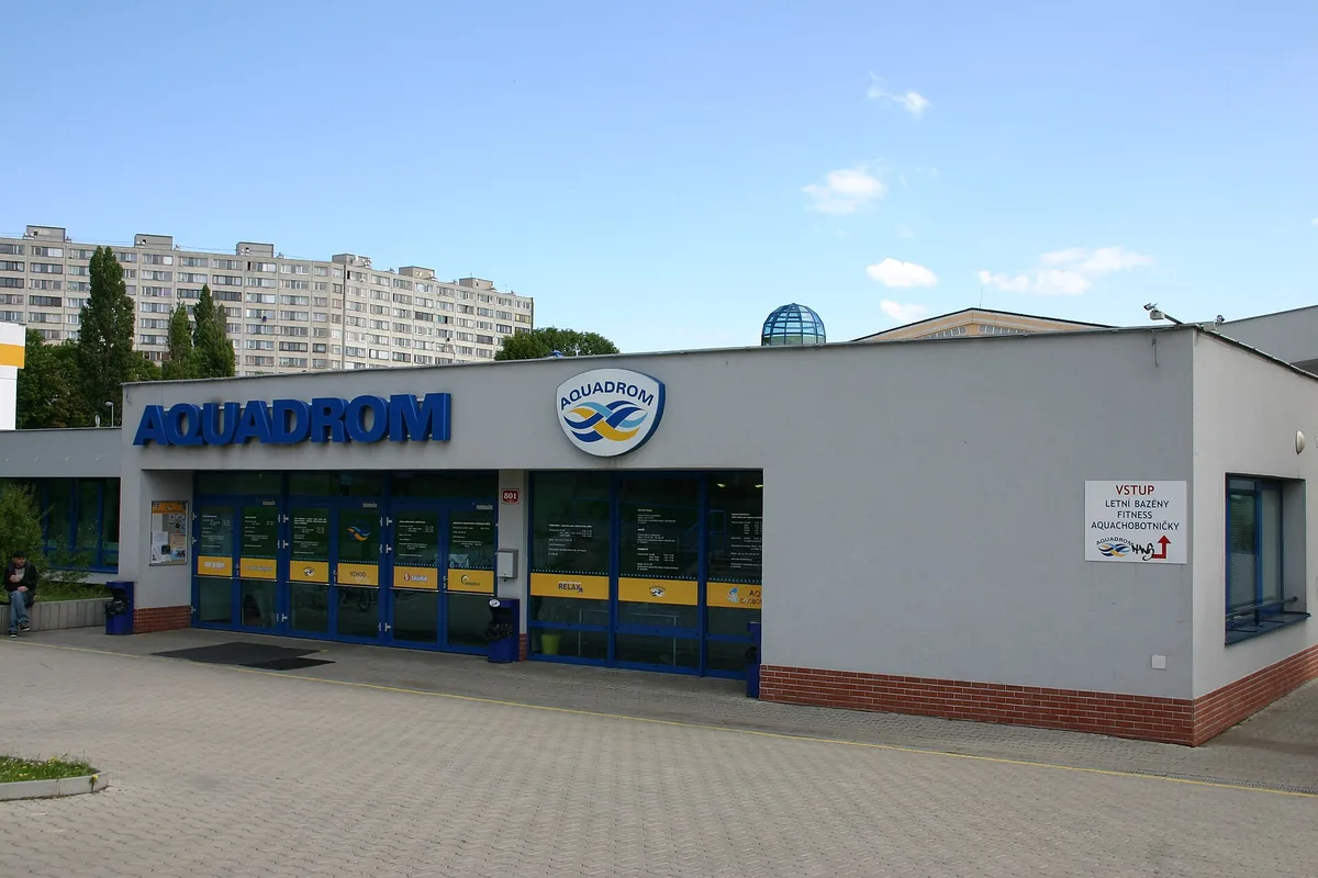 Šedá přízemní budova Aquadromu Most s modrým nápisem Aquadrom a prosklenými vstupními dveřmi, v pozadí panelový dům