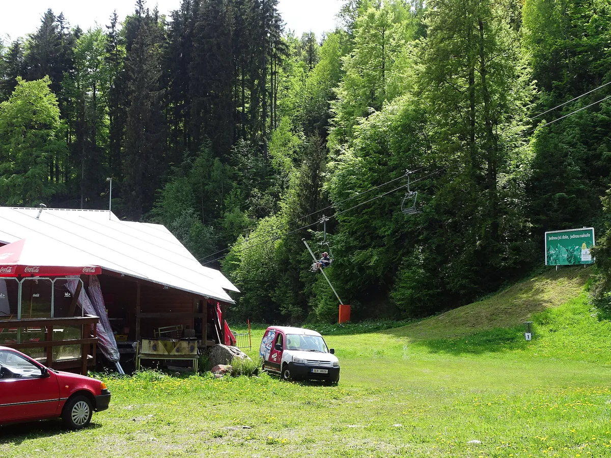 Sedačková lanovka na Medvědín ve Špindlerově Mlýně nad loukou u lesa, v popředí občerstvení a zaparkovaná auta