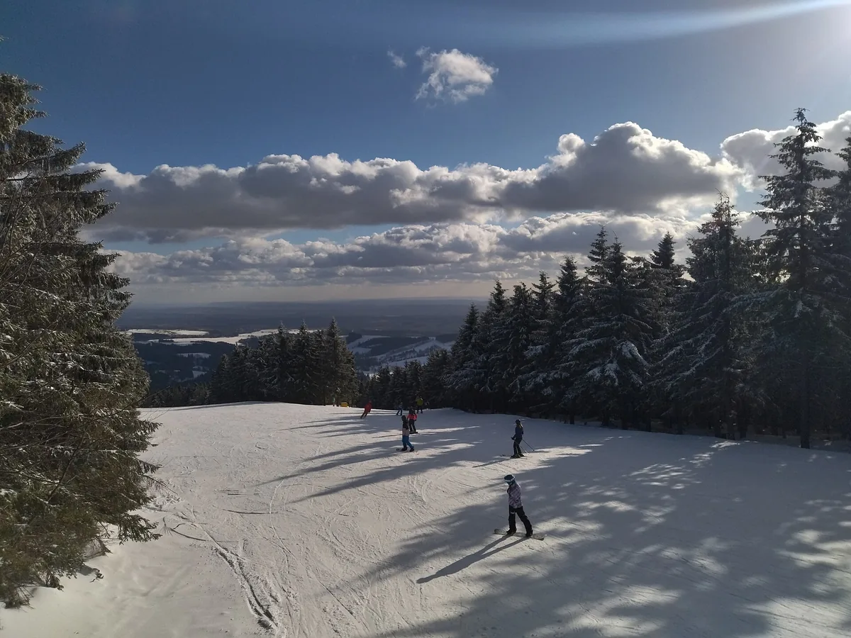 Zasněžená sjezdovka lemovaná smrky ve Ski centru Říčky v Orlických horách s lyžaři a výhledem do podhůří