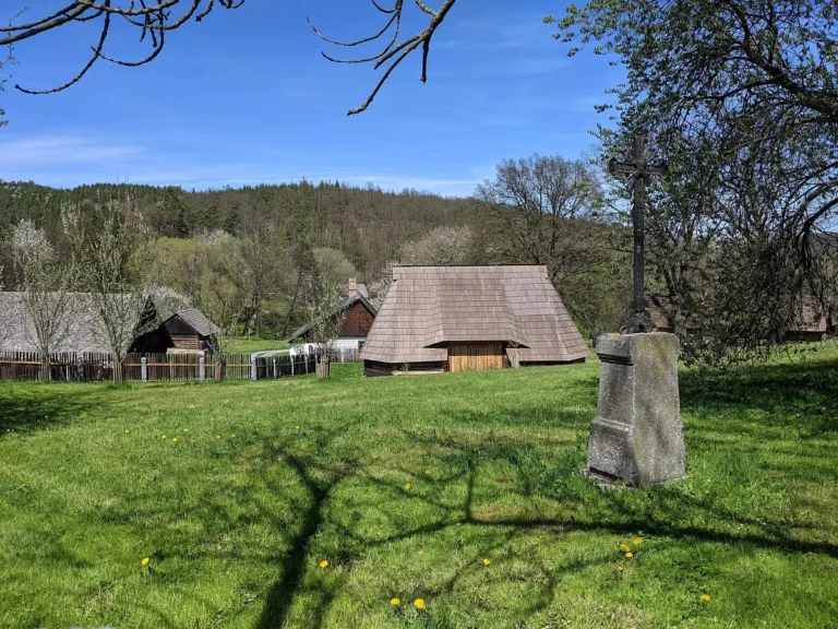 Louka ve skanzenu Vysoký Chlumec s kamenným soklem kříže a roubenými stavbami se šindelovými střechami pod zalesněným svahem