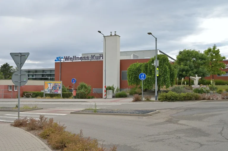Cihlově červená budova Wellness Kuřim s nápisem na fasádě a vysokým komínem, pohled přes křižovatku v červenci 2026