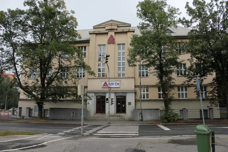 Budova pobočky Všeobecné zdravotní pojišťovny v Karviné s logem nad vchodem, před ní přechod pro chodce a stromy